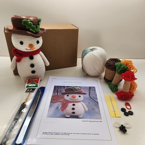 May include: A crochet Christmas snowman kit with white yarn, red, green, and orange yarn, a crochet hook, a needle, and a pattern. The kit includes instructions for making a snowman with a red scarf and green holly.