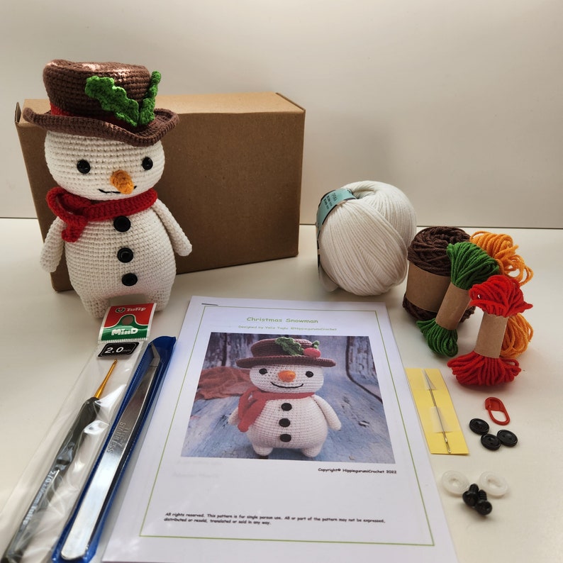 May include: A crochet Christmas snowman kit with white yarn, red, green, and orange yarn, a crochet hook, a needle, and a pattern. The kit includes instructions for making a snowman with a red scarf and green holly.