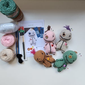 Könnte beinhalten: Ein Set aus vier gehäkelten Voodoo-Puppen mit verschiedenen Garnfarben und Knöpfen für die Augen. Die Puppen haben Stecknadeln, die in sie gesteckt sind, und sind auf einer weißen Oberfläche angeordnet. Die Puppen werden mit Garn, Nadeln und einer Anleitung zum Herstellen von Voodoo-Puppen geliefert.