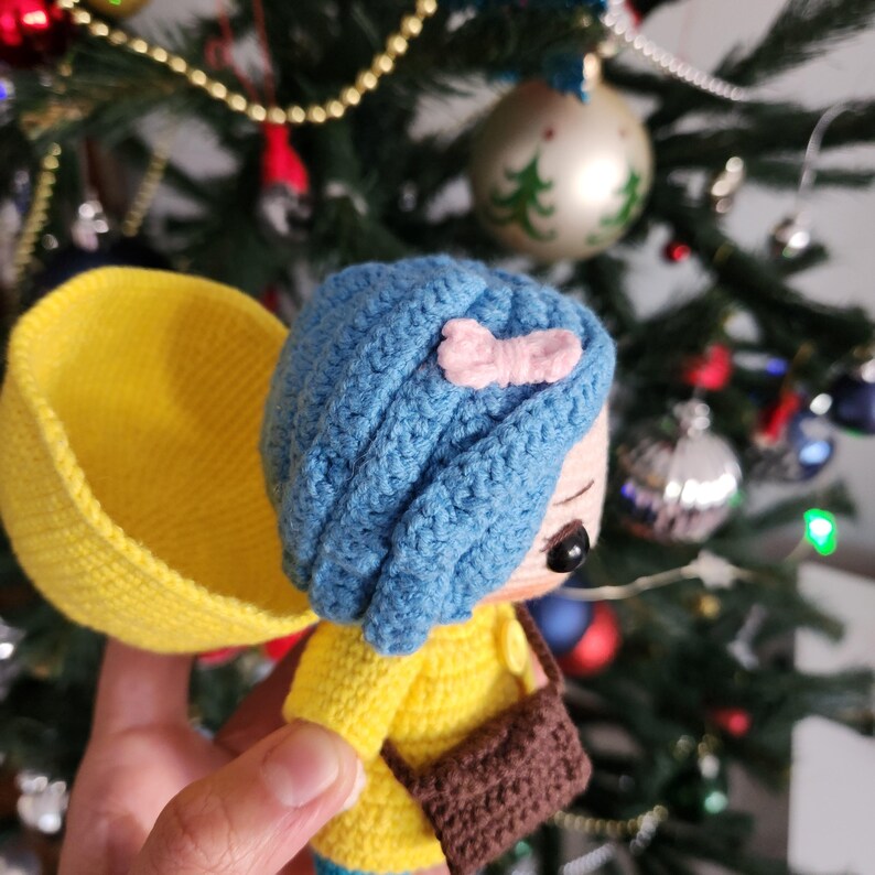 May include: A handmade crochet doll with blue hair, a pink bow, and a yellow jacket. The doll has a brown purse and is holding a yellow object. The background is a Christmas tree with ornaments.