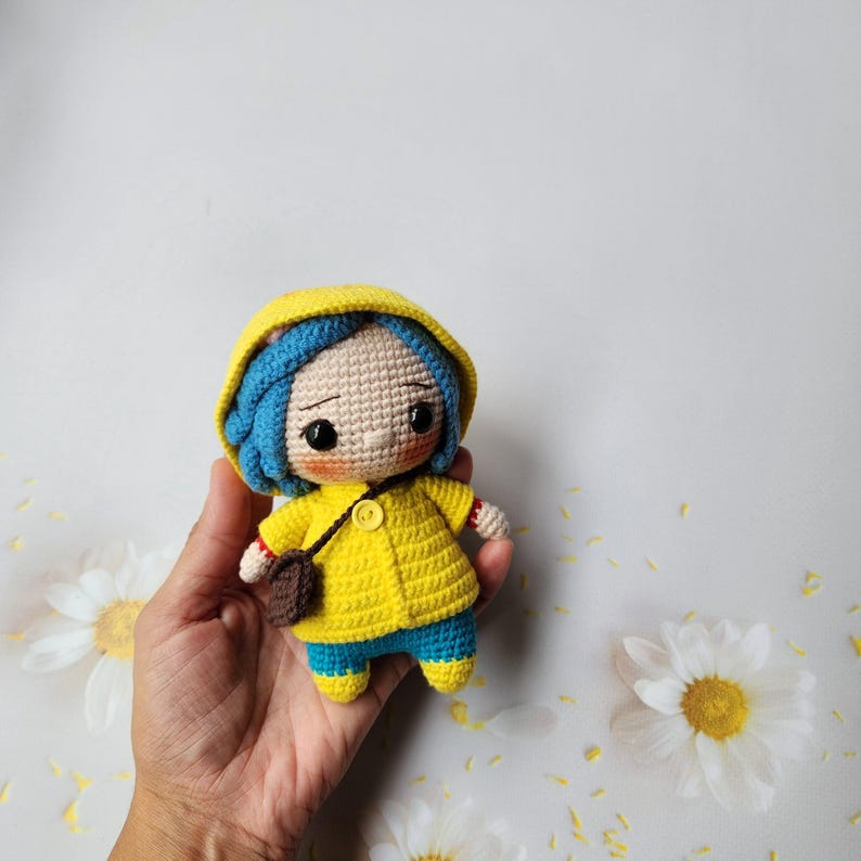 May include: Crocheted doll of a character wearing a yellow hooded jacket, blue pants, and a brown satchel. The doll has blue hair and a neutral expression. The doll is handmade and has a button closure on the jacket.