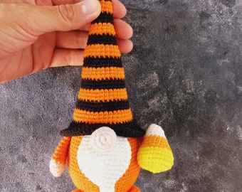 May include: A crocheted gnome wearing an orange and black striped hat and holding a yellow and orange candy corn.