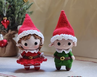 May include: Two crocheted elf ornaments, one in red and white with a brown wig and the other in green and white. Both have a white trim around their hats and are standing on a white and brown surface.