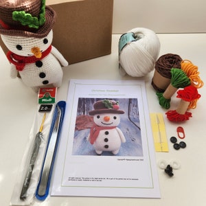 May include: A crochet Christmas snowman kit with white yarn, green, red, and orange yarn scraps, a crochet hook, a pair of tweezers, a needle, safety pins, and black and white buttons. The kit also includes a pattern for crocheting a snowman.