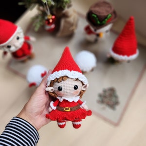 May include: A crocheted elf doll with a red hat, white trim, and a red dress. The elf has brown hair and brown eyes. The doll is holding a small red bag.