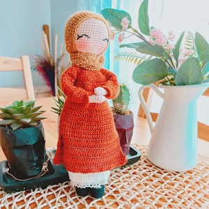 May include: A crocheted doll wearing a long orange dress with a white lace trim and a brown headscarf. The doll has a smiling face with closed eyes and is standing on a woven mat.