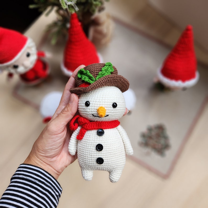 May include: A white crocheted snowman with a brown hat, green leaves, and a red scarf. The snowman has black eyes, a carrot nose, and black buttons.
