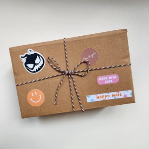 May include: A brown cardboard box with a green and red string tied around it. The box has several stickers on it, including a black and white pumpkin, a pink sticker that says "Made with Love", a brown sticker that says "Thank you", and a colorful sticker that says "Happy Mail".