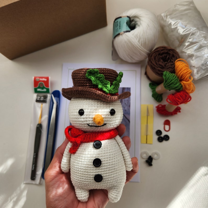 May include: A crocheted snowman toy with a brown hat, green holly leaves, and a red scarf. The snowman has black eyes, a carrot nose, and three black buttons.