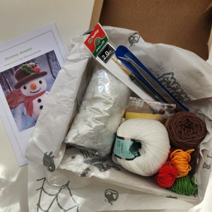 May include: A craft kit for crocheting a Christmas snowman. The kit includes white yarn, brown yarn, orange yarn, green yarn, stuffing, a crochet hook, a pair of scissors, and instructions. The kit also includes a pattern for a crocheted snowman.