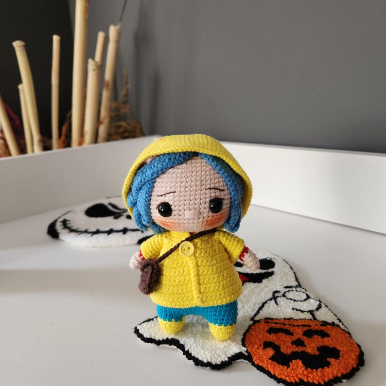 May include: A handmade crochet doll with blue hair, wearing a yellow hooded jacket, blue pants, and a brown satchel. The doll is standing on a white rug with a ghost and jack-o'-lantern design. The doll has a button closure on the jacket.