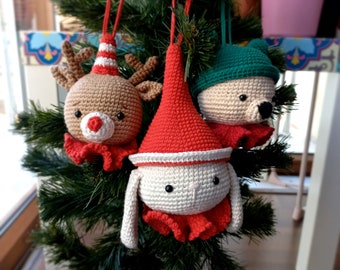 May include: Three crocheted Christmas ornaments hanging on a green Christmas tree. The ornaments are a reindeer, a bunny, and a bear. The reindeer is brown with red and white accents. The bunny is white with red and white accents. The bear is brown with green and white accents.