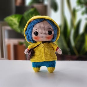 May include: A handmade crochet doll featuring a yellow hooded jacket, blue pants, and blue hair. The doll has a small brown satchel and is approximately 6 inches tall. The doll's face has black eyes and rosy cheeks.