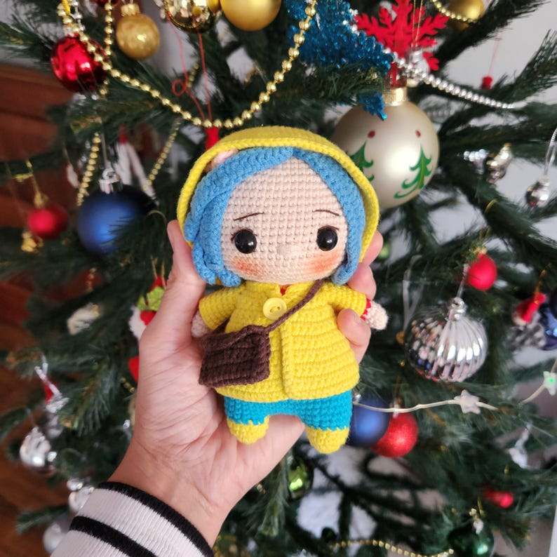 May include: A handmade crochet doll featuring a character with blue hair, wearing a yellow coat, blue pants, and a brown satchel. The doll has large, expressive eyes and is approximately 6 inches tall. The doll is a unique, handcrafted item.