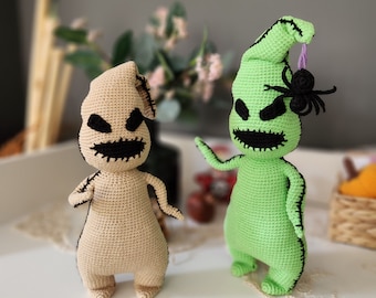 May include: Two crocheted plush toys, one green and one beige, are posed on a white surface. The green toy is shaped like a ghost with a black stitched face and a purple hat. The beige toy is shaped like a ghost with a black stitched face and a pointed hat.
