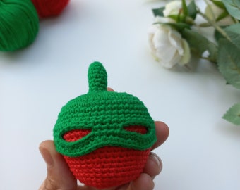 May include: A crocheted red and green superhero toy with a green mask. The toy is being held in a hand.