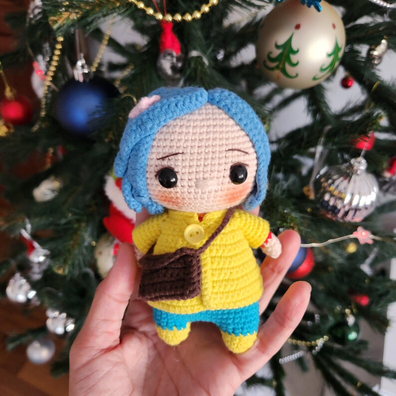 May include: Crocheted doll with blue hair, wearing a yellow jacket, blue pants, and a brown bag. The doll has large, expressive eyes and a small pink flower in its hair. The background is a Christmas tree with ornaments.