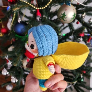May include: A handmade crochet doll with blue hair, a yellow jacket, and a yellow basket. The doll has a brown strap and blue pants. The doll is set against a Christmas tree background.