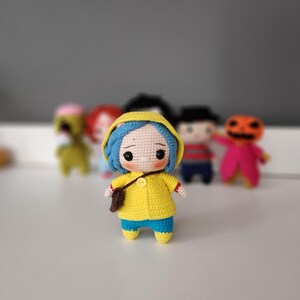 May include: A handmade crochet doll with blue hair, wearing a yellow raincoat and blue pants. The doll has a small brown bag and is standing in front of other crochet dolls. The doll has a sad expression.