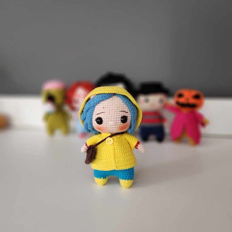 May include: A handmade crochet doll with blue hair, wearing a yellow raincoat and blue pants. The doll has a small brown bag and is standing in front of other crochet dolls. The doll has a sad expression.