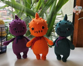 May include: Three crocheted plush toys in the shape of monsters. The monsters are purple, orange, and green. Each monster has a spiky head, large eyes, and a small mouth.