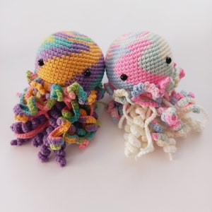 Può includere: Due giocattoli a forma di medusa all'uncinetto. Uno è di colore arcobaleno con tentacoli viola e l'altro è di colore rosa pastello e blu con tentacoli bianchi.