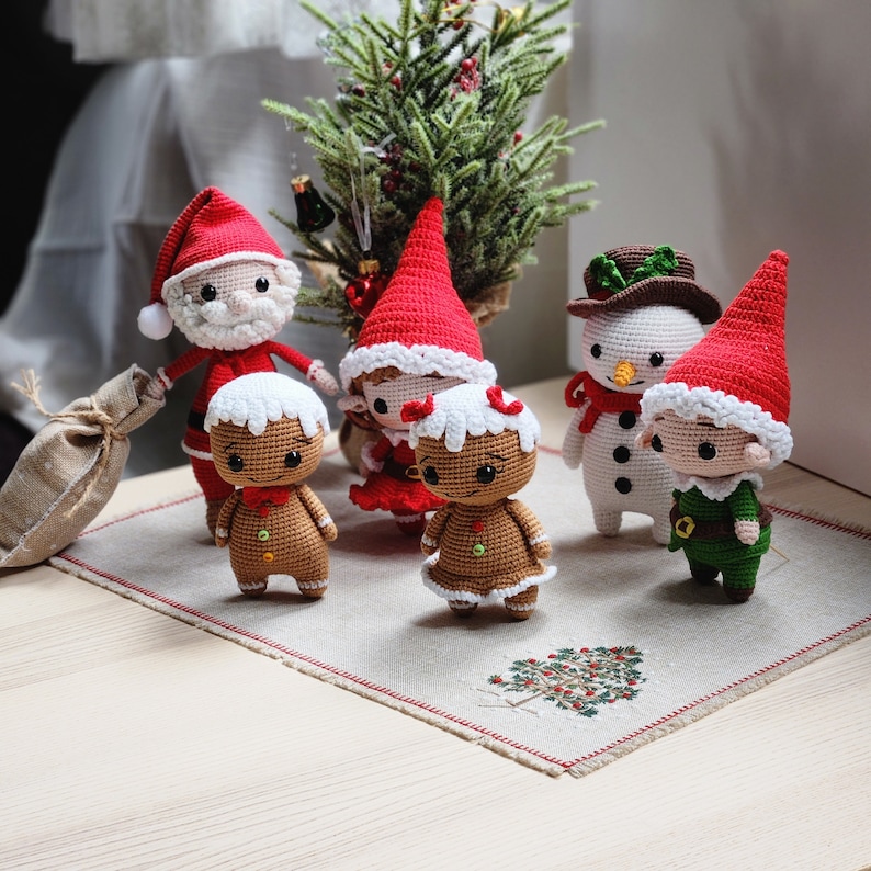 May include: A collection of crocheted Christmas decorations, including a gingerbread person, a snowman, a Santa Claus, and two elves. The decorations are arranged on a white tablecloth with a red and white border.