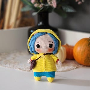 May include: A handmade crochet doll with blue hair, wearing a yellow hooded jacket, blue pants, and yellow shoes. The doll has a small brown satchel and is standing on a white surface. The doll is a whimsical, handcrafted item.