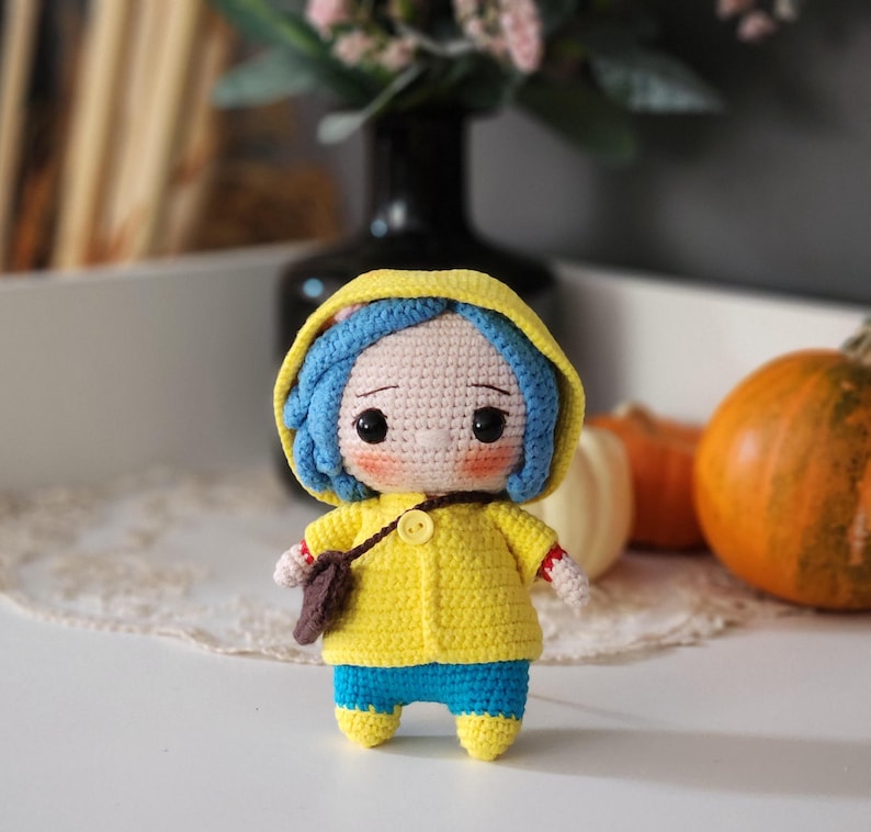 May include: A handmade crochet doll with blue hair, wearing a yellow hooded jacket, blue pants, and yellow shoes. The doll has a small brown satchel and is standing on a white surface. The doll is a whimsical, handcrafted item.
