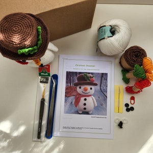 May include: A crochet kit for making a Christmas snowman. The kit includes white yarn, brown, green, red, and orange yarn scraps, a crochet hook, a needle, buttons, safety eyes, and a pattern. The pattern is titled "Christmas Snowman" and is designed by "Helga Tighe".