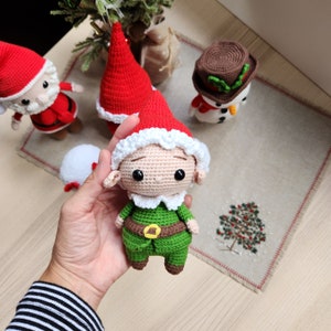 May include: A crocheted elf doll wearing a green outfit, white trim, and a red Santa hat. The elf has a brown belt and a brown face with black eyes.