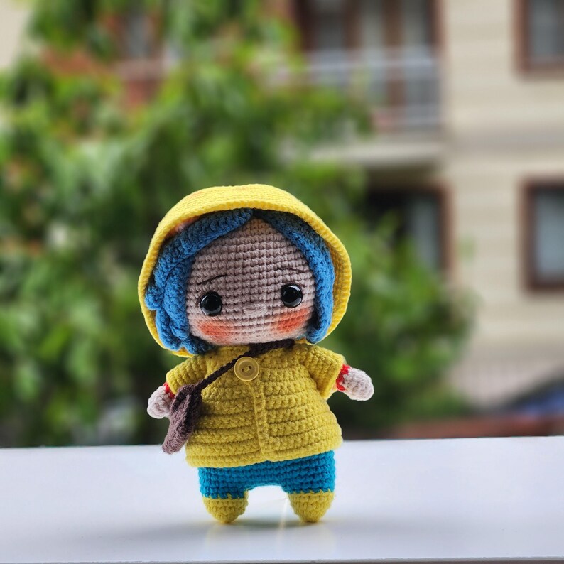 May include: A handmade crochet doll with blue hair, wearing a yellow hooded jacket, blue pants, and a brown satchel. The doll has large black eyes and a button closure on the jacket. The doll is approximately 6 inches tall.