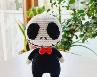 May include: A crocheted plush toy resembling a skeleton wearing a black suit and a red bow tie. The toy has white skin, black eyes, and a stitched-on smile.