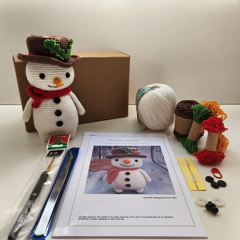 May include: A crochet Christmas snowman kit with white yarn, red, green, and orange embroidery floss, a brown hat, black buttons, and a crochet hook. The kit also includes a pattern for making the snowman.