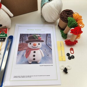 May include: A crochet kit for making a snowman. The kit includes white yarn, brown yarn, red yarn, green yarn, orange yarn, black buttons, safety eyes, a crochet hook, a needle, and a pattern. The pattern is titled "Christmas Snowman" and includes instructions for making the snowman.