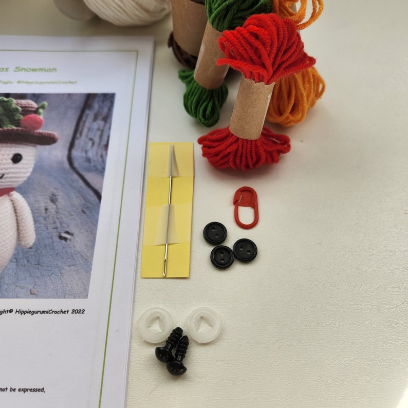 May include: A crochet kit for making a Christmas snowman. The kit includes white, red, green, and orange yarn, a needle, safety eyes, buttons, and a plastic safety pin. The kit also includes a printed pattern.