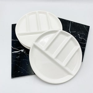 May include: Two white ceramic divided plates. Each plate has a circular shape with three rectangular compartments and a larger semi-circular section. The plates are stacked on a black and white marble-patterned surface.