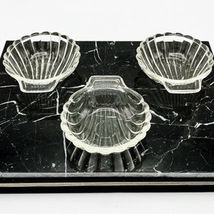 May include: Three clear glass bowls shaped like seashells on a black marble tray.