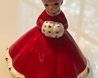 Könnte beinhalten: Eine Keramikfigur einer Person in einem roten Kleid und einer passenden roten Weihnachtsmütze mit weißem Pelzbesatz mit schwarzen Punkten. Die Figur hat blondes Haar und hält einen weißen Pelzmuff mit schwarzen Punkten vor der Brust.