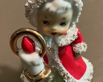 May include: A ceramic figurine of a child wearing a red coat with white fur trim and a green skirt. The figure wears a matching red hood with white fur trim and holds a white candle with a gold handle and a red flame in one hand.