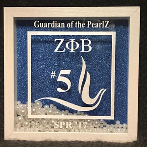 May include: A white framed shadow box with a blue glitter background. The box contains white pearls and a white vinyl design that says "ZΦB #5" and "SPR '17" with a white wave design. The text "Guardian of the PearlZ" is at the top of the box.