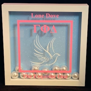May include: A white framed shadow box with a blue background and white glitter. The box has a pink border and the letters "GΦΔ" in pink. A white dove is flying in the center of the box. The words "Lone Dove" and "The Exquisite One" are written in pink at the top and bottom of the box. There are white pearl beads around the edge of the box.