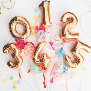May include: Gold foil number balloons, including 0, 1, 2, 3, 4, and 5, are arranged against a white background. Each balloon is adorned with colorful tissue paper and ribbons. Confetti in various colors adds a festive touch.