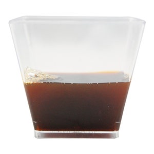 May include: A clear, square-shaped plastic cup filled with a dark brown liquid. The liquid is about halfway up the cup and has a slightly foamy surface. The cup is set against a plain white background.