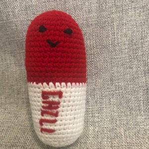 May include: A red and white crocheted pill-shaped toy with a smiling face and the word "Happy" written on the white part.