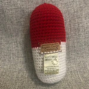 May include: A red and white crocheted pill-shaped toy with a brown label that reads "Handcrafted in the USA by Bugzie's Crochet Creations" and "BUGZIE'S CROCHET CREATIONS bugziescrochet etsy.com Handmade in Smithville, MO, USA of domestic & imported materials".