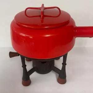 May include: A bright red enameled fondue pot with a matching lid featuring a cross-shaped handle. The pot is supported by a dark brown metal stand with three legs, each ending in a rounded foot, and includes a central burner component underneath.