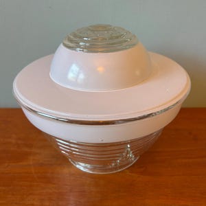 May include: A vintage light fixture with a clear glass shade and a white, saucer-shaped base. The glass shade has horizontal ridges, and the fixture has a retro, mid-century modern aesthetic. The fixture is sitting on a wooden surface.