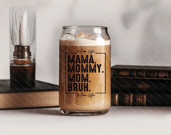 May include: A glass can with a black and white graphic that reads "Mom life MAMA. MOMMY. MOM. BRUH. Mom life". The can is filled with a brown liquid.