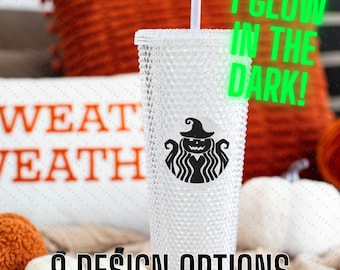 May include: A clear, textured, reusable cup with a black witch design and the text "I GLOW IN THE DARK!"  The cup is sitting on a white surface with a pillow and pumpkins in the background.